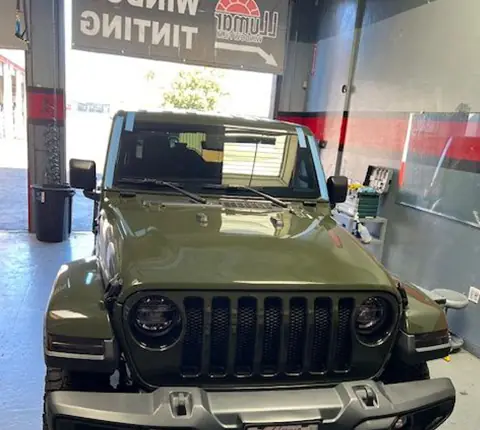 An olive Jeep Wrangler parked under the Window Tinting sign inside the A1 Auto Glass bay, Llumar banner overhead